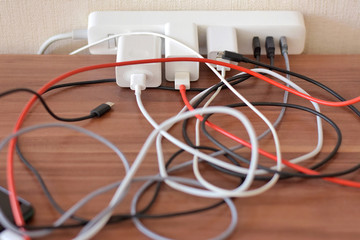 Blurred tangled colorful electricity, usb cables on wooden table. White socket set with mess connection wires for charging gadgets with selective focus. Electrical power outlet 