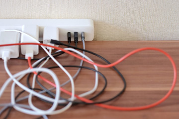 Blurred tangled colorful electricity, usb cables on wooden table. White socket set with mess connection wires for charging gadgets with selective focus. Electrical power outlet 