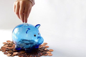Child`s hand putting money in blue transparent piggy bank on pennies with copy space