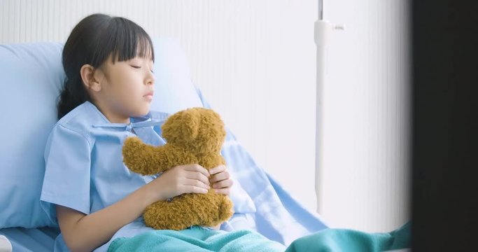 Little asian girl lying alone in hospital and hugging toy, unhappy child missing parents, illness and treatment.