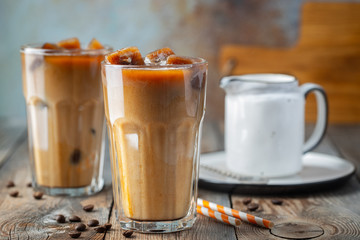 Ice coffee in a tall glass with cream poured over, coffee ice cubes and beans on a old rustic wooden table. Cold summer drink with tubes on a blue rusty background with copy space