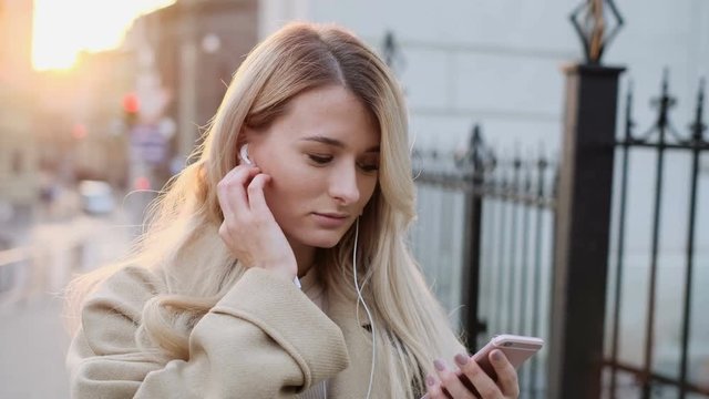 Smiling girl in headphones using smartphone, listening to music and browsing on smartphone. Outdoor portrait. Blogger scrolls through social media on device, changing tracks and reading news on app