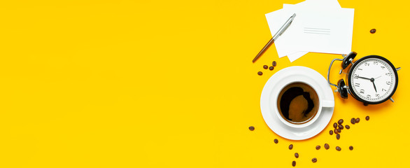 Flat lay cup black coffee, coffee beans, black alarm clock, pens, white cards on yellow background...