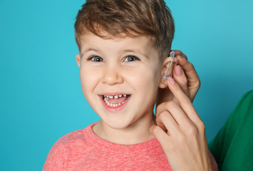 Young woman adjusting little son's hearing aid on color background, closeup