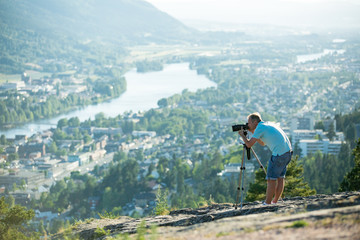 Fototapeta na wymiar Man filming on camera with tripod on the top of mountain. Tourist hiking and taking pictures. Panoramic top view of Drammen city, mountains in fog and valley with river, sunny weather. Norway