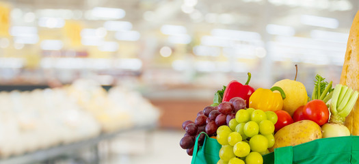 Fresh fruits and vegetables in reusable green shopping bag with supermarket grocery store blurred defocused background with bokeh light