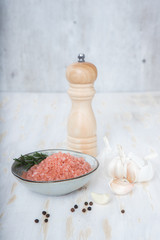 A sprig of rosemary lies in a white bowl with pink Himalayan salt, near other spices and a pepper mill.
