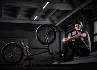 Young Bmx rider relaxing after practicing tricks with his bike in a skatepark indoors