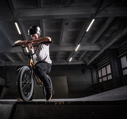 Fototapeta na wymiar Tired BMX rider in protective helmet sitting on his bicycle in a skatepark indoors