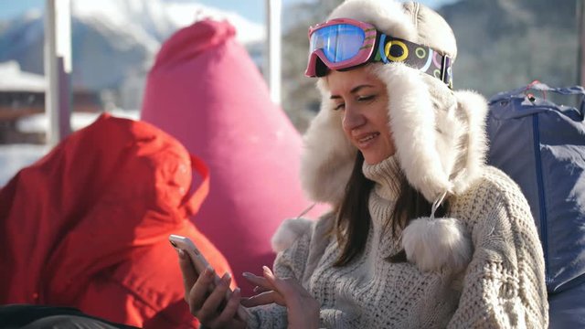 A girl uses a smart phone sitting in a cafe at a ski resort. Young woman relaxing at an alpine ski resort.