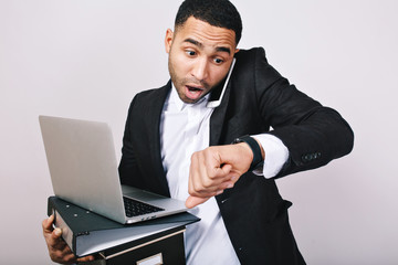 Busy handsome guy in white shirt and black jacket talking on phone, holding folders, laptop, looks astonished at watch on white background. Being late, meeting, office worker, career