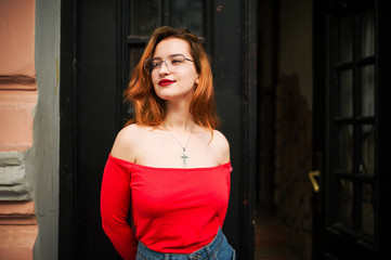 Attractive redhaired woman in eyeglasses, wear on red blouse and jeans skirt posing at street against old wooden door.