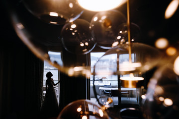 Look from behind a shiny lamp at thoughtful bride standing before the window
