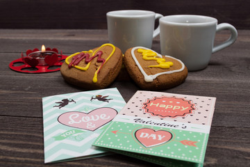 Two white cups of coffee, cookies in the shape of a heart, red candle and a Valentine card on a wooden table on Valentine's Day