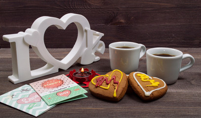 Two white cups of coffee, cookies in the shape of a heart, red candle and a Valentine card on a wooden table on Valentine's Day