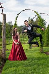 Beautiful teen girl in red gown smiling at joyful teen boy in tuxedo jumping in beautiful park setting © Lisa Kim