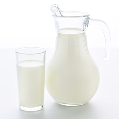 Transparent jug and a glass completely filled with fresh cow milk stand on a clean white background.