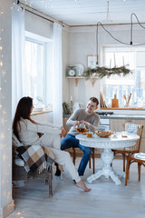 Young caucasian couple sitting at home kitchen, hugging, smiling, drinking tea. Young lovers having good time. Man and woman communicate sitting at a table in the dining room.Winter decor.