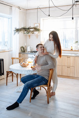 Young caucasian couple sitting at home kitchen, hugging, smiling, drinking tea. Young lovers having good time. Man and woman communicate sitting at a table in the dining room.Winter decor.
