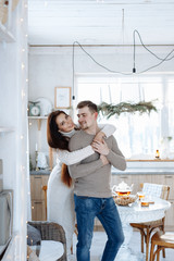 Young caucasian couple sitting at home kitchen, hugging, smiling, drinking tea. Young lovers having good time. Man and woman communicate sitting at a table in the dining room.Winter decor.