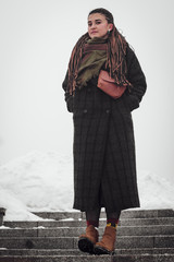 stylish photograph of a portrait of an attractive woman with long pigtails, dressed in an eco-coat and scarf, walked in a winter-frozen city