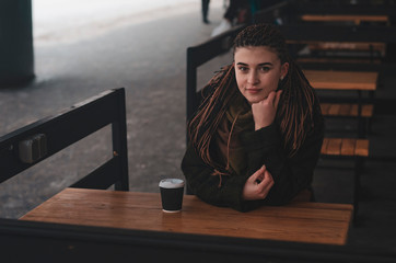 stylish portrait of an attractive woman with long pigtails, in an eco-coat, scarf, sitting at a table outdoors holding coffee cup of hot coffee
