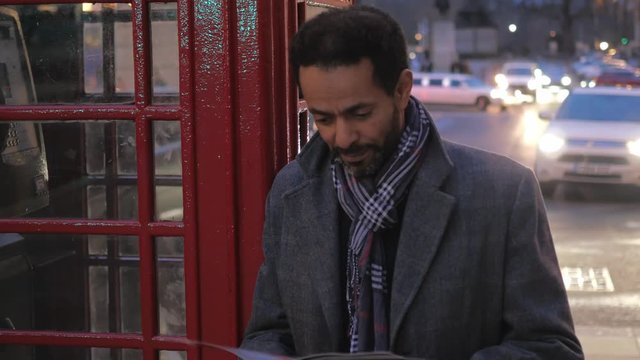 Man in the city of London reads a magazine while leaning against a phone booth