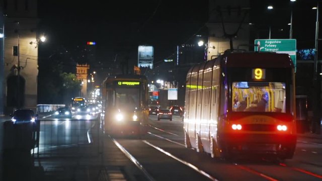 Night view of the yellow tram approaching and stopping with cars passing-by on the busy road in modern city. Traffic in the night city.