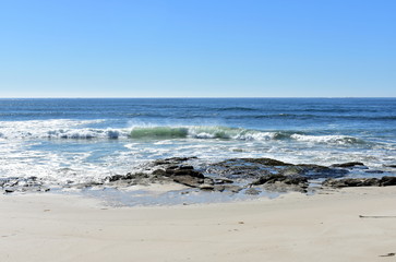 Fototapeta na wymiar Beach with rocks, bright sand and small waves breaking. Blue sea with foam, sunny day. Galicia, Spain.
