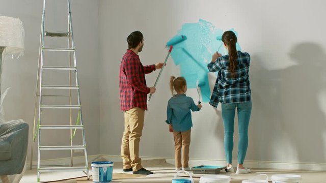 Family Time Together with Small Daughter. Young Father and Mother Showing Their Child How to Paint a Wall with a Roller. Paint Color is Light Blue. Room at Home is Prepared for Renovations.