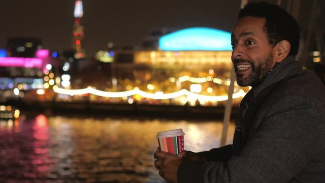 Man drinks coffee on a bridge in London and enjoys the city lights at night
