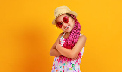 Funny happy child girl in summer dress, pink glasses and hat is laughing on yellow background