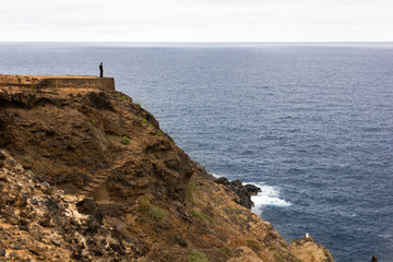 Fototapeta na wymiar Man standing on cliff edge by the sea. Person alone on top of mountain facing the ocean on cloudy day. Loneliness, solitude, depression concepts