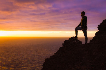Visionary man standing on top of cliff edge staring at colorful sunset by the sea in Gran Canaria. Silhouette of person witnessing unique twilight from mountain top. Successful, entrepreneur concepts - Powered by Adobe