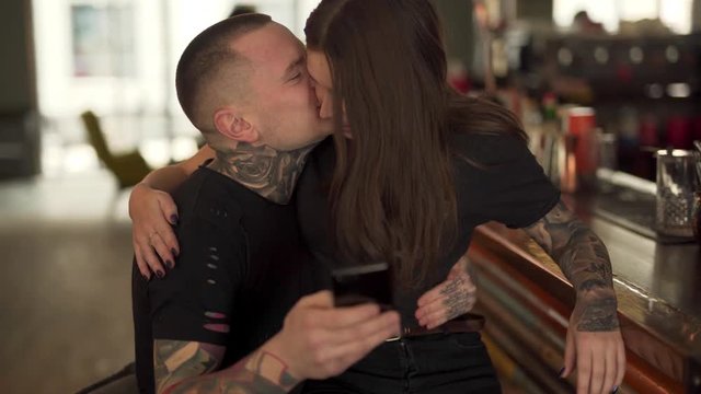Cute tattooed couple is sitting in a cafe, clicking on the phone, hugging and kissing each other. Man and woman has a lot of tattoos on their hands and necks and are wearing a black t-shirts.
