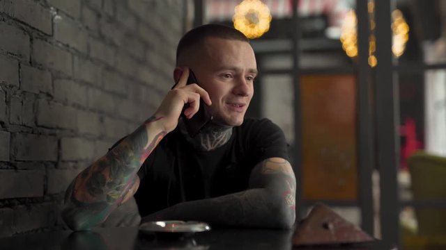 A handsome man fully covered in tattoos talks on his phone. He is happy and just makes small talks. The background is blurred.