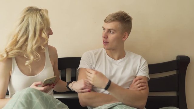 Couple dressed in white T-shirts lying in bed covered with blanket. Blond girl touching boyfriend's arm, smiling and asking him to take photo/selfie together while making puppy-dog eyes