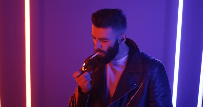 Caucasian stylish good looking man in leather black coat and with beard lighting a cigarette and smoking on the blue and pink neon light wall background.