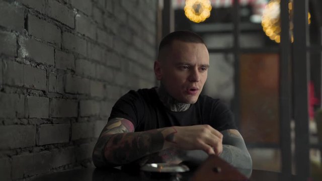 A handsome man fully covered in tattoos smokes and enjoys it to the great degree. He is thoughtful and kind of stressed in the beginning, but smiles in the long run.  The background is blurred.