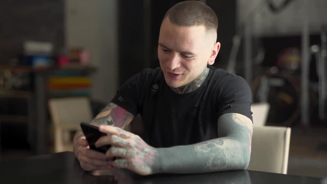 A handsome man fully covered in tattoos chews a gum and has a video call via his phone. He is happy and smiles during the whole conversation. The background is blurred.