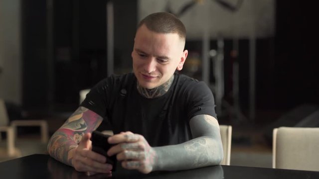 A handsome man fully covered in tattoos is busy on his phone. He also smiles and tells amusing stories.