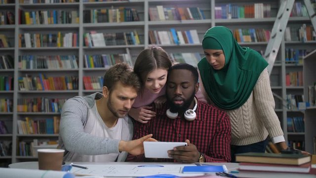 Group of excited mulit ethnic college students checking exam grades online on digital tablet in university library. Joyful diverse high school friends with tablet pc celebrating passing final exam.