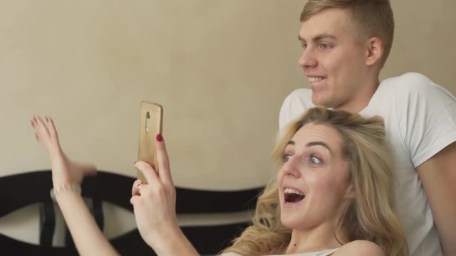 Boy and girl dressed in white T-shirts. Beautiful blond girl lying on the bed trying to make selfie with her handsome boyfriend. The boy kissing her on forehead. Lovers/couple concept