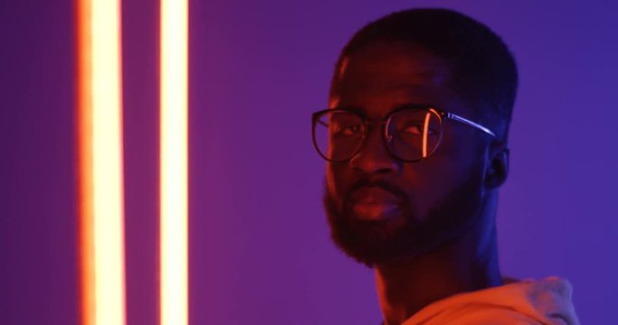 Close upof teh profile of the young African American man in glasses turning his head to the camera and smiling. Portrait. Neon lights.