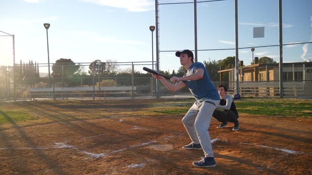 A young man baseball player hits a bunt with his bat and runs to first base during a team practice in the park at sunset SLOW MOTION.