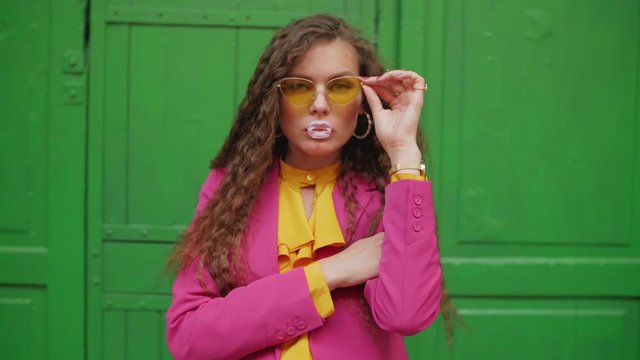 Young fashionable woman chewing bubble gum, blowing bubblegum bubble, looking at camera. Model wearing trendy yellow sunglasses, pink blazer
