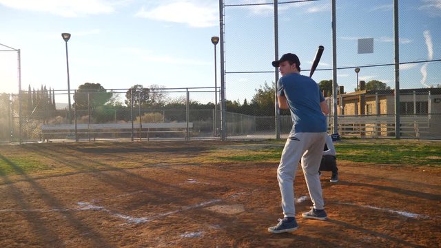 A young man baseball player hits a home run in slow motion and runs to first base during a team practice in the park at sunset.