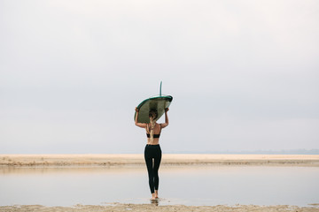 Female surfer wearing dreadlocks hair style carrying long board on her head going to the sea