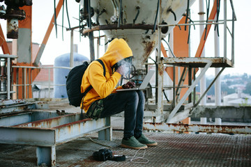 Hacker or man wearing yellow hoody with laptop seating in abandoned industrial building, smoke and use computer