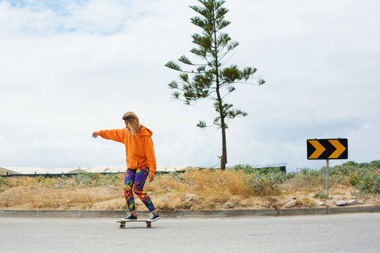 Street portrait of positive young female wearing orange hoody riding with skateboard in city